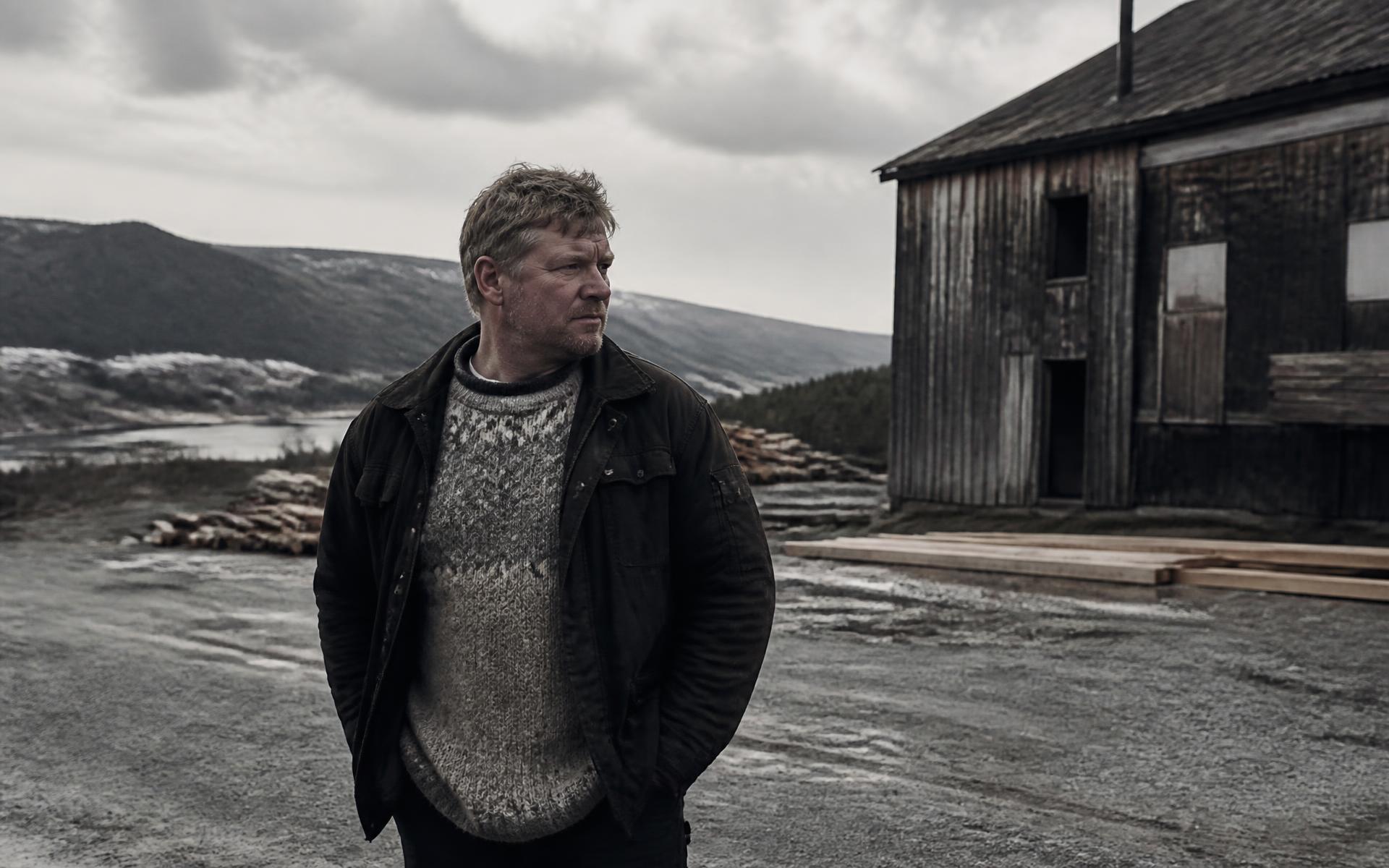 Mann i arbeidsklær foran et gammelt trelastbygg i et norsk elvelandskap